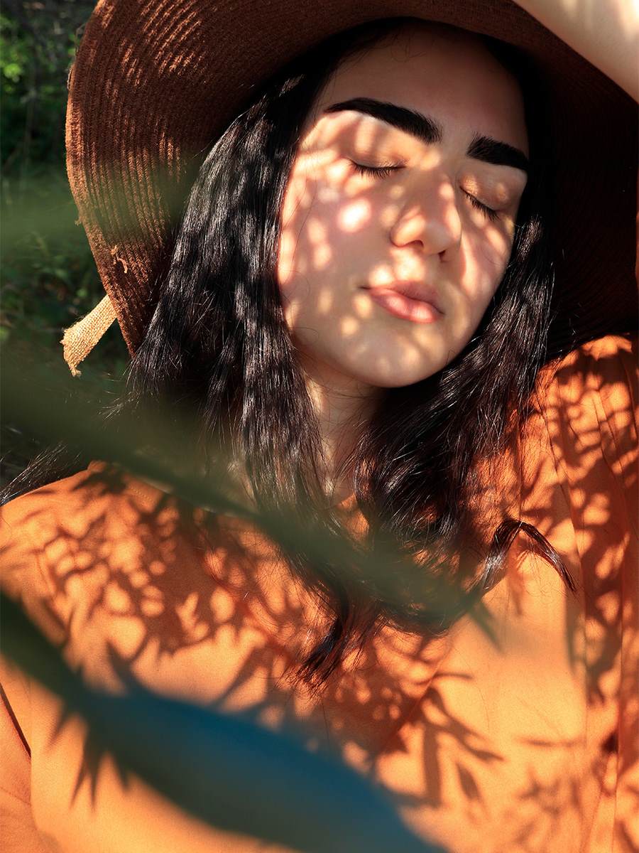 Hispanic woman with long straight black hair wearing a hat. She has her eyes closed and is enjoying the shade on a sunny day.
