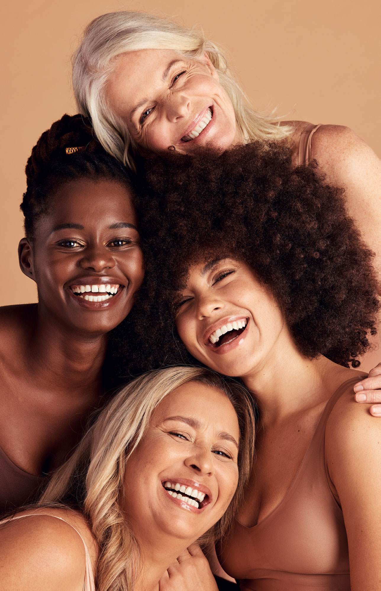4 smiling women. An older white women with blonde hair. A young black woman with black hair. A young woman with light brown skin and brown hair in a natural style. A young white woman with blonde hair.