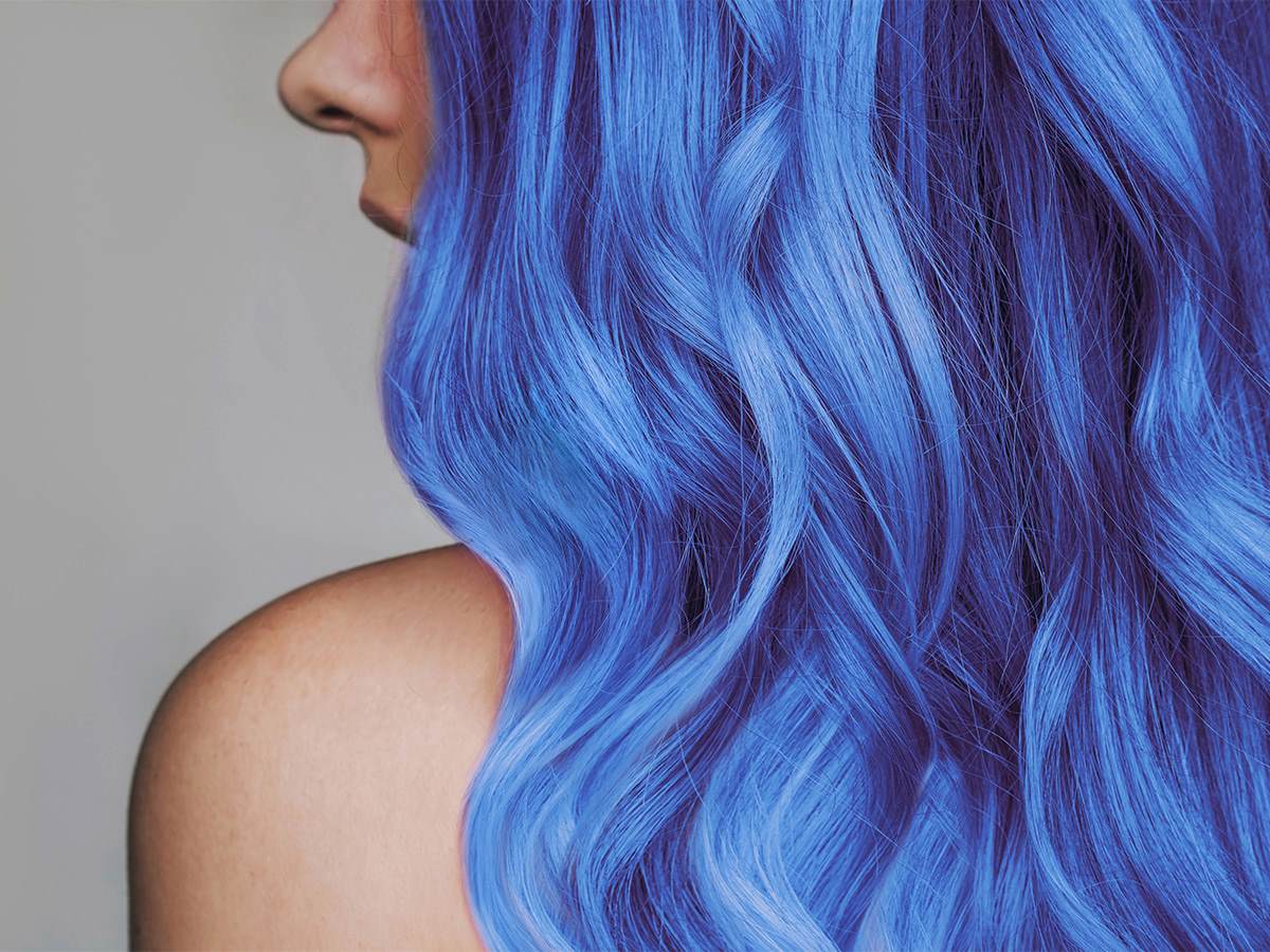 Photo showing a white woman from behind where all we can see is her hair, shoulder and nose. Her hair is blue, long and wavy.