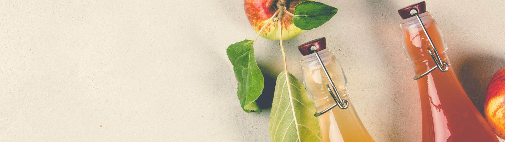 Cropped top of 2 glass bottles containing a light brown and brownish red liquid respectively. The liquid is apple cider vinegar. There is a red and green apple next to them with its leaves attached.