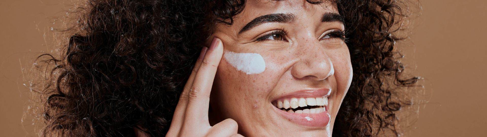 The face of a smiling woman in her twenties with long, very curly, brown hair. Her skin is light brown in colour with noticeable freckles. She is smiling and looking up and to the right. With two fingers of her right hand she is applying a white cream to her face.