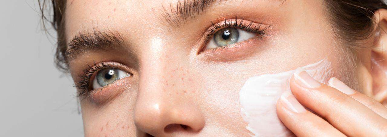 Close up on the nose and green eyes of a young white woman looking up and to the left and applying a white cream to her check with her left hand.