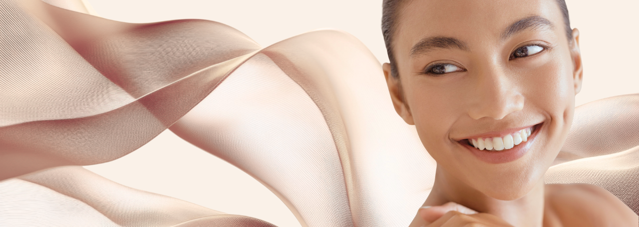 A wide, panoramic composition centers on a smiling young woman whose face occupies the right side of the frame. Her skin is smooth and softly illuminated by warm, diffused light that brings out a natural glow on her cheekbones and the curve of her jaw.