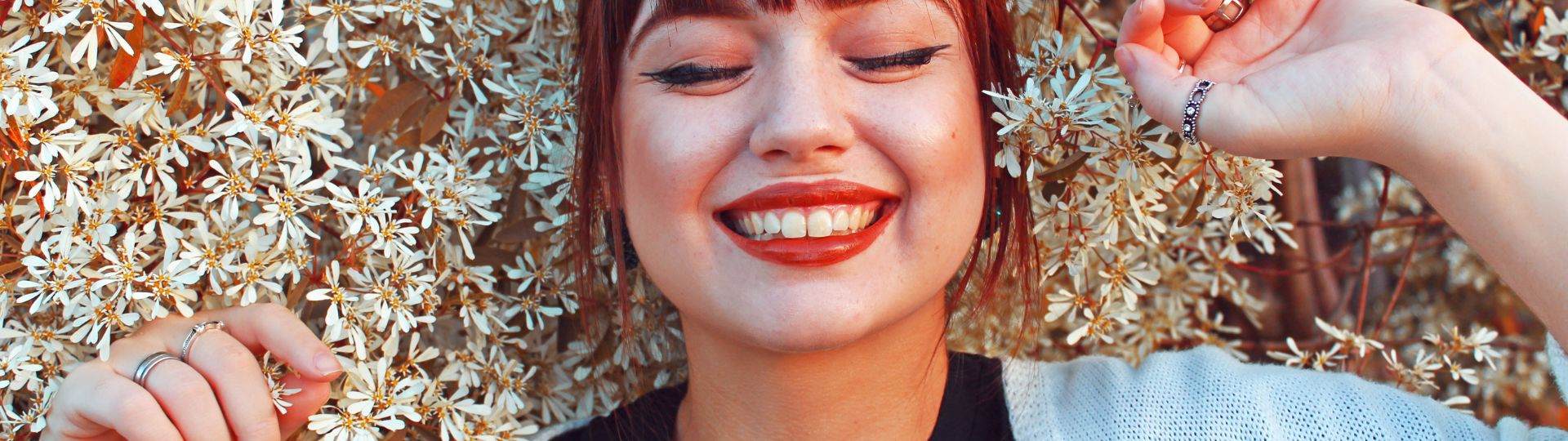 Woman smiling with her eyes closed. She has white skin and dyed red hair. She is standing amongst dozens of white flowers which drape around her.