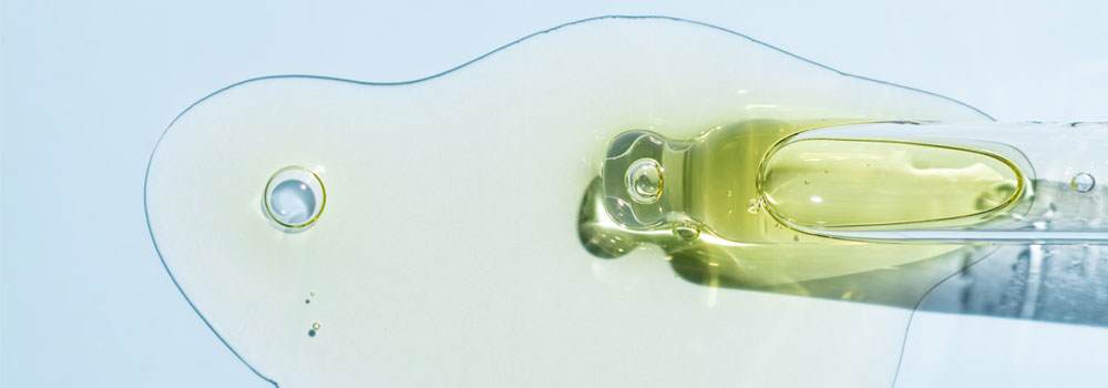 Close up on the end of a glass pipette that is filled with a green/yellow liquid. The liquid is spilling out onto a white surface.