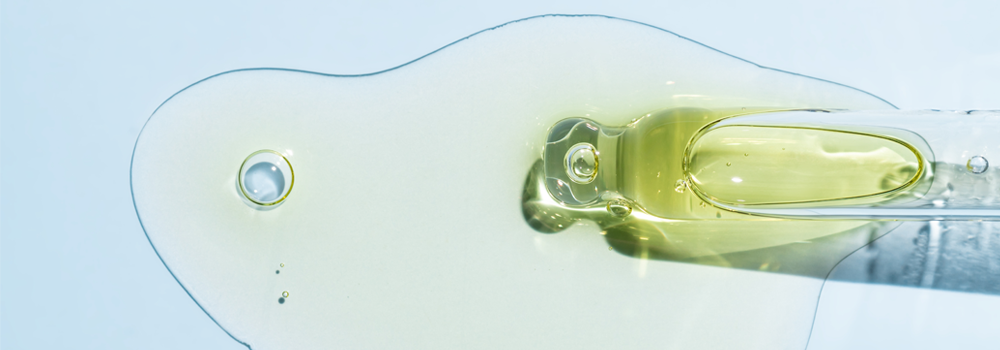 Close up on the end of a glass pipette that is filled with a green/yellow liquid. The liquid is spilling out onto a white surface.