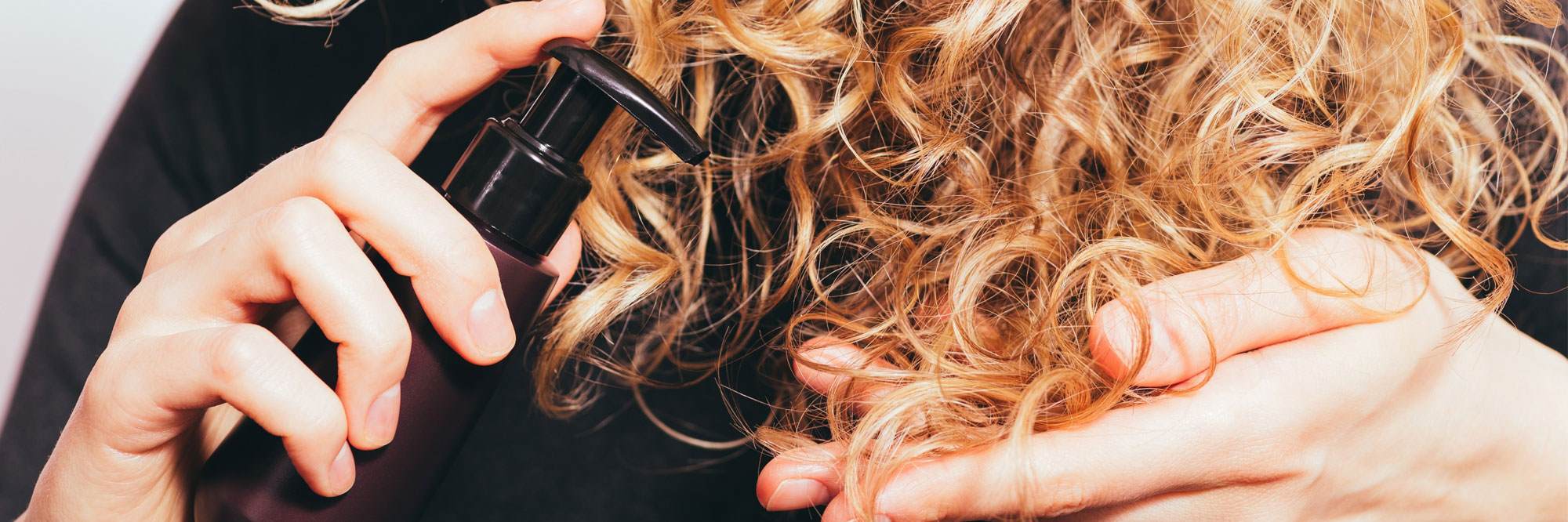 Cropped photo of the hands of a young white woman applying hair serum to her curly blonde hair