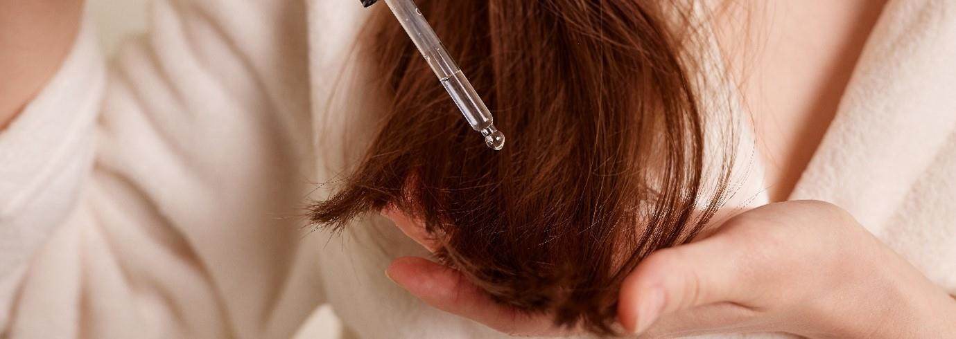 A cropped photo showing a white woman with reddish-brown straight hair applying a serum to her hair.