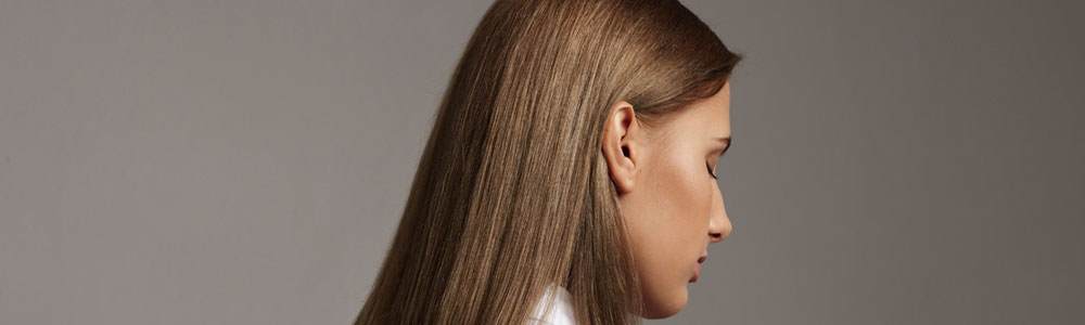 Photo of a young white woman with long straight light brown hair. She is photographed from behind with her hair in focus, her face is looking away from us and to the right. She has her eyes closed. Her hair is glossy.