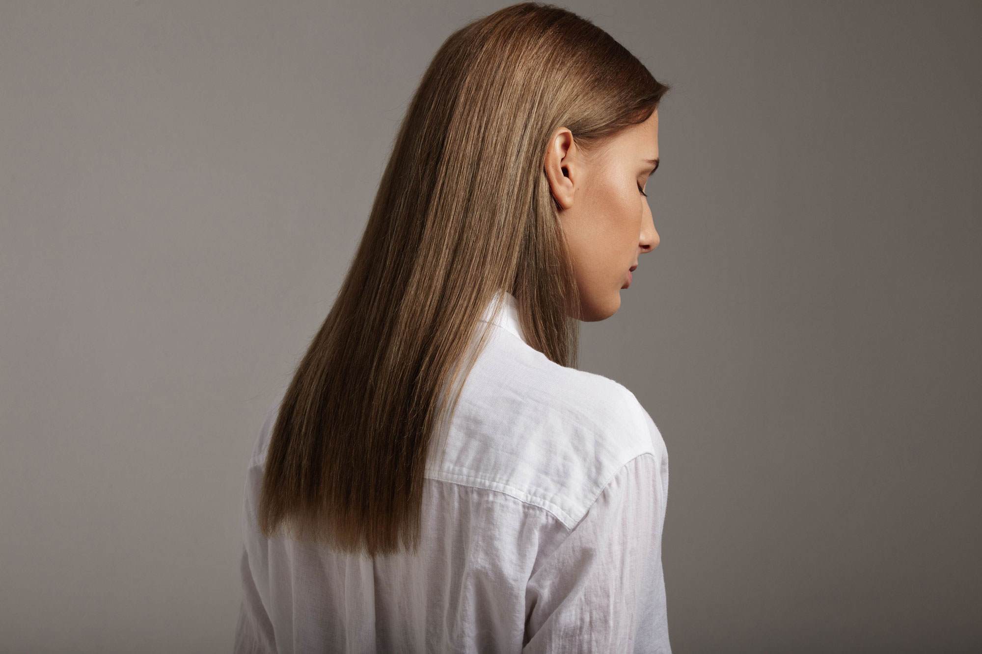 Photo of a young white woman with long straight light brown hair. She is photographed from behind with her hair in focus, her face is looking away from us and to the right. She has her eyes closed. Her hair is glossy.