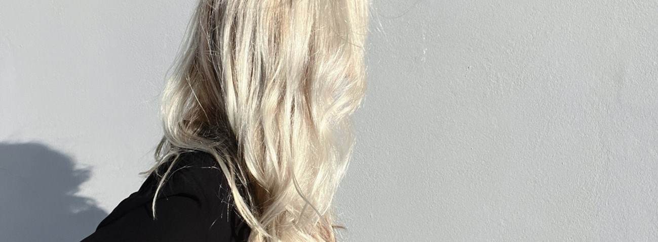 A photo of a woman with blonde hair whose face is hidden beneath her hair. The focus of the image is her hair which is shiny, wavy and healthy.