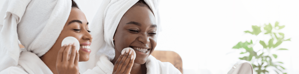 Mild and gentle - Smiling black female friends testing beauty products at home, using cotton pads, looking at mirror, bedroom interior, sun flare. 