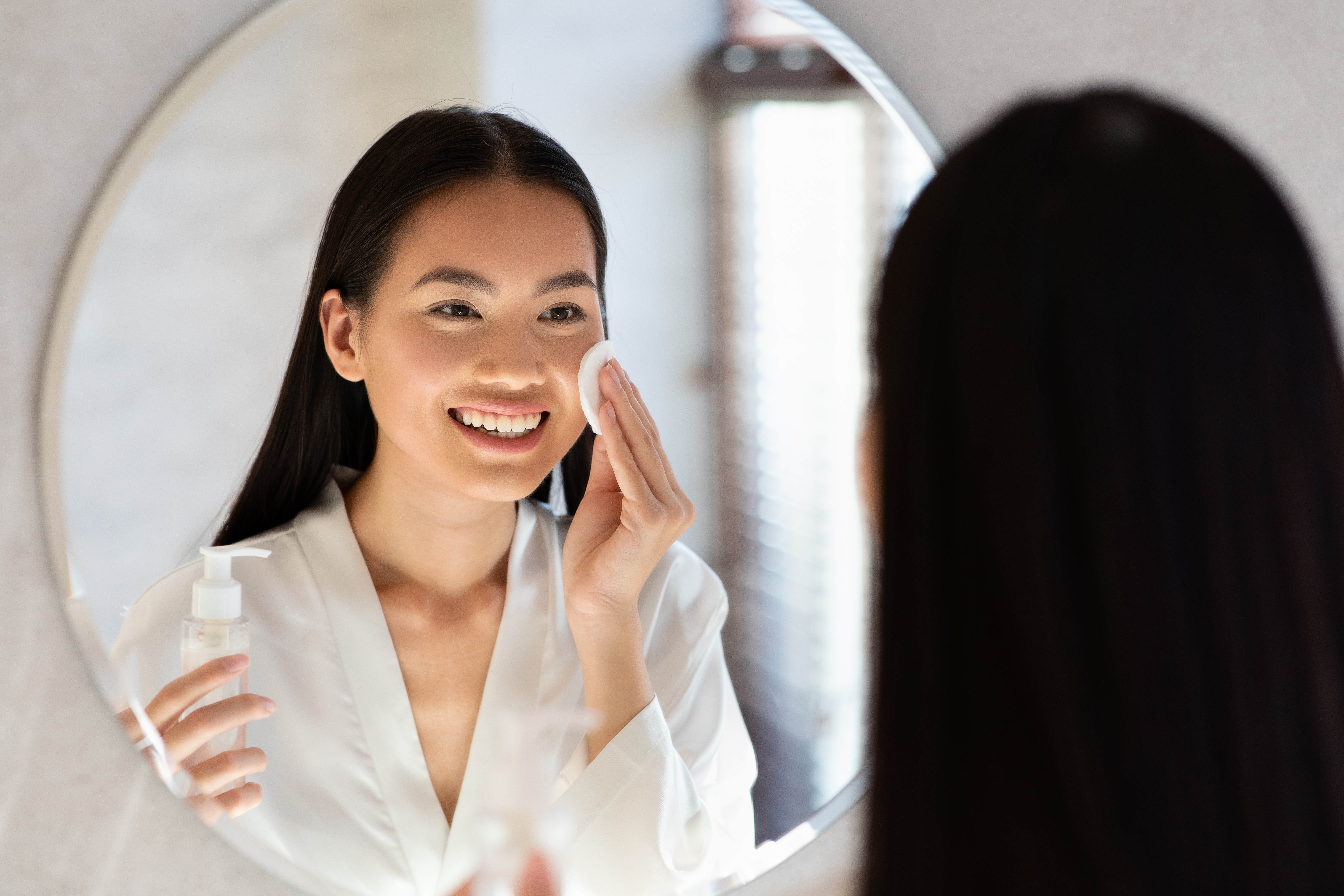 asian lady in bathrobe using cleansing milk, removing makeup in front of mirror at bathroom, woman using facial tonner and cotton pad, over shoulder photo
