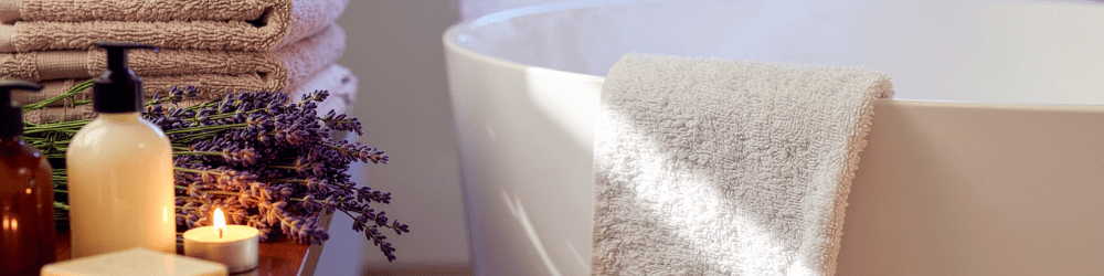 Cleansing for self-care – Bathroom scene with towel over bath, pile of towels, lavender, candle and bottle of cleanser