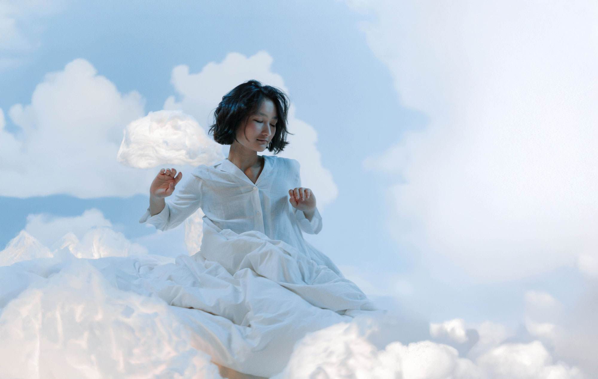 A young east asian woman with black hair cut into a short bob. She is wearing white pyjamas and covered in a white duvet. Her eyes are closed and arms are posed as if just waking up. She is surrounded by white clouds as if she is in the sky.