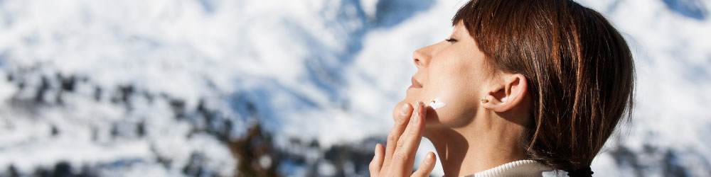white woman with brunette hair applying sun cream in front of mountains with snow while sun is shining