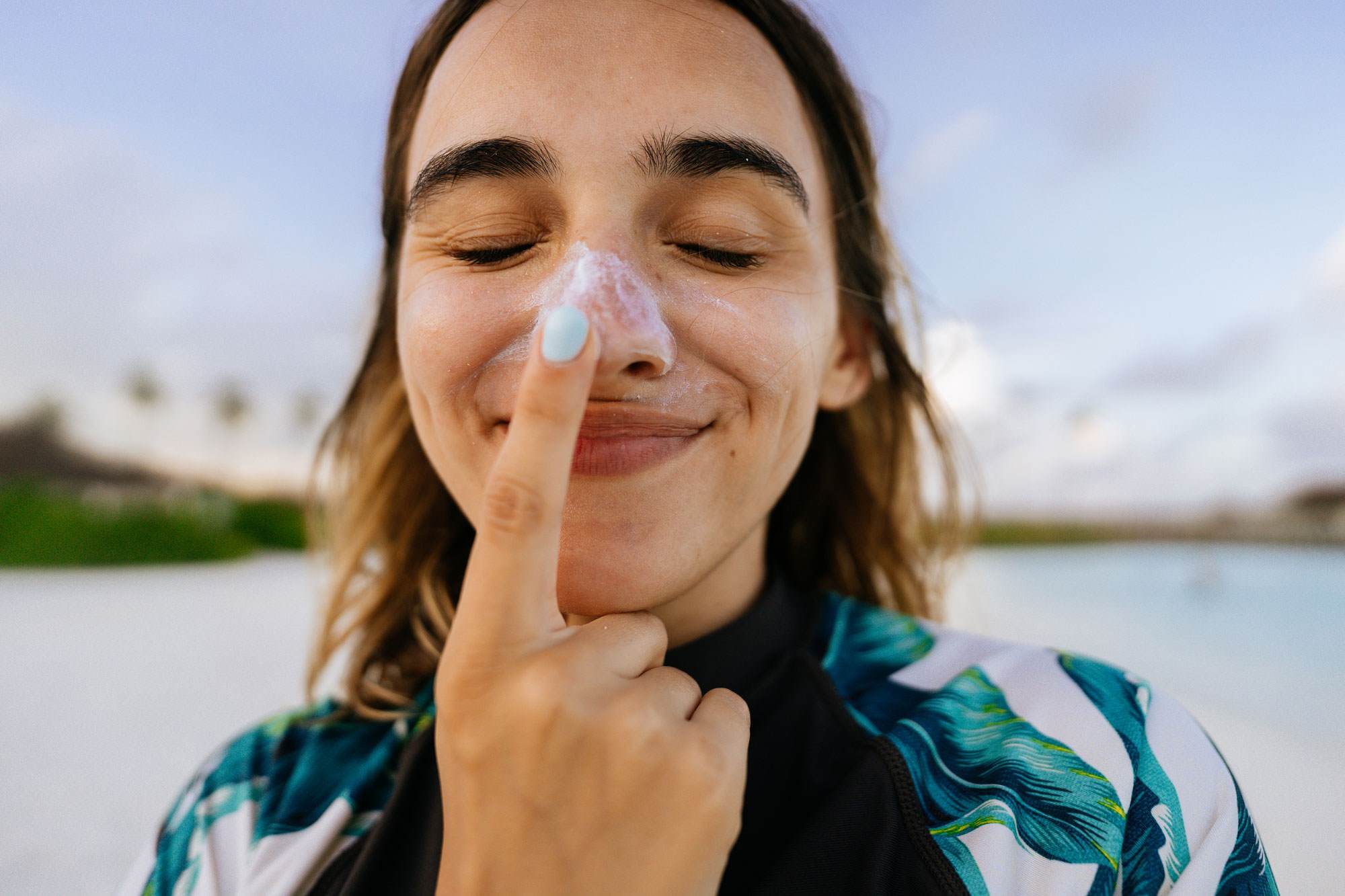 Young white woman applying sunscreen while being outdoors, at the beach; making silly faces while applying SPF and protecting her skin from the sun. 