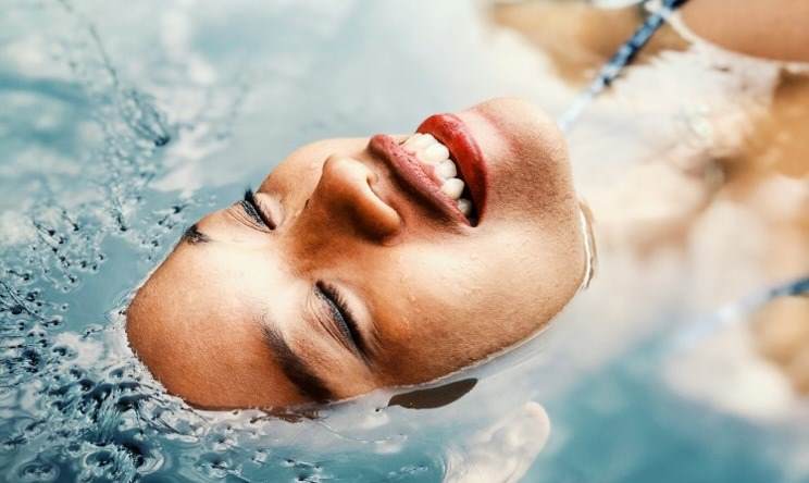 Close up on the face of a woman floating in a pool of water. She ahs her eyes closed and her mouth opened smiling. She has light brown skin.