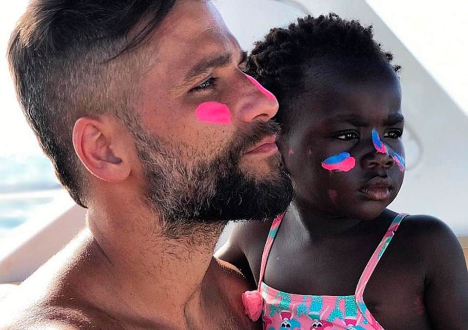 A father and daughter. Both are looking to the right. The father has white skin and brown to black straight hair with a beard. He is holding his daughter, a little girl with black skin and curly black hair. Both are looking to the right. Both are wearing brightly coloured pink and blue sunscreen.