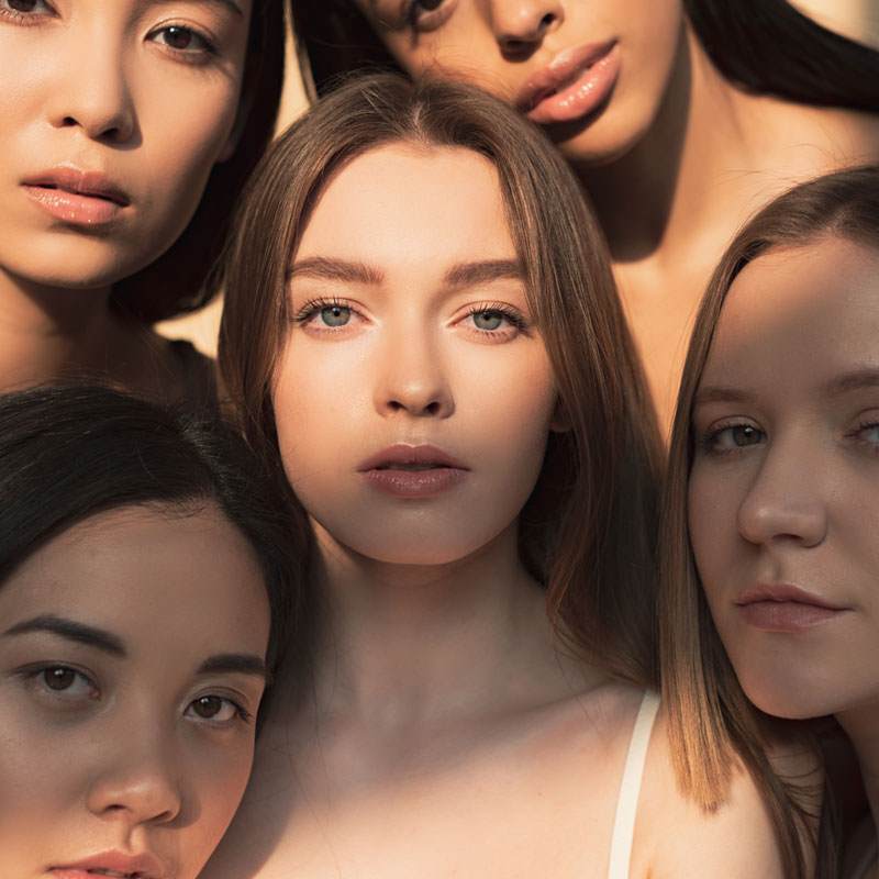 5 young women with different hair colours and ethnicities looking directly at the camera. The are clustered close together.