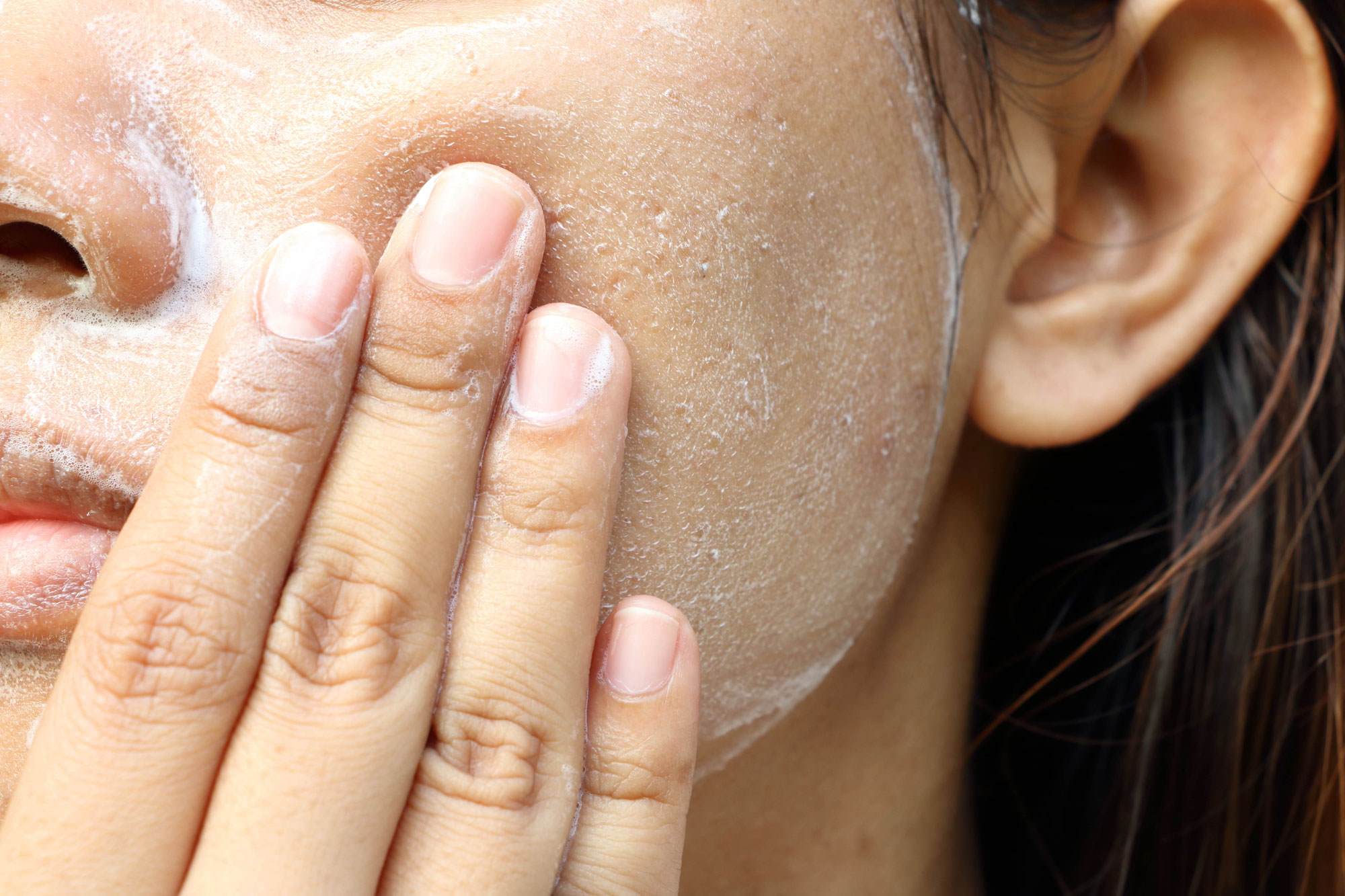 Close up of one side of a White woman’s face. She is applying foaming cleansing to her cheek with visible foam covering her cheek, finger tips, and nose.