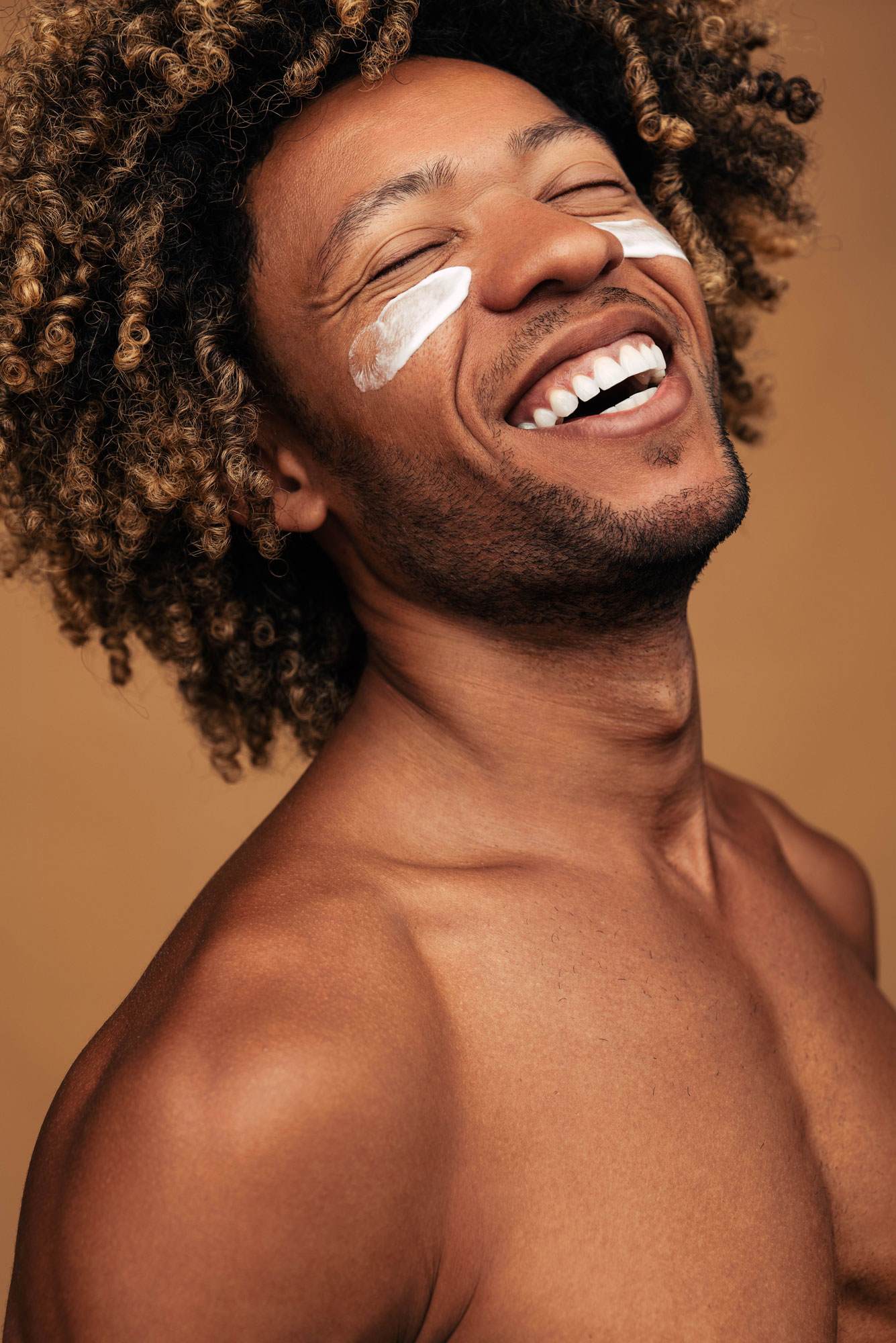 Portrait of Black male, showing his shoulders, chest and full face and large afro hair style. His head is tilted upwards and he is smiling, showing teeth and with eyes closed. He has face cream applied to his cheeks just under eyes, in two nearly symmetrical lines.