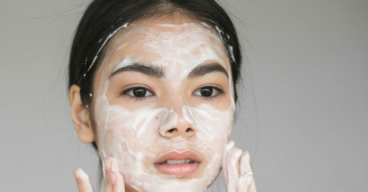 Younger East Asian woman with straight black hair applying a soft cleansing cream with French rose to her face. 