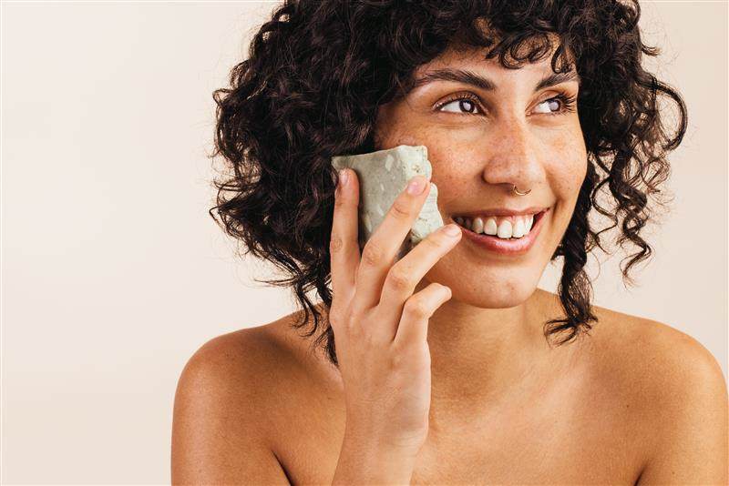 Young white woman with brown curly hair and a nose piercing holding a green solid format cleansing bar to her face.
