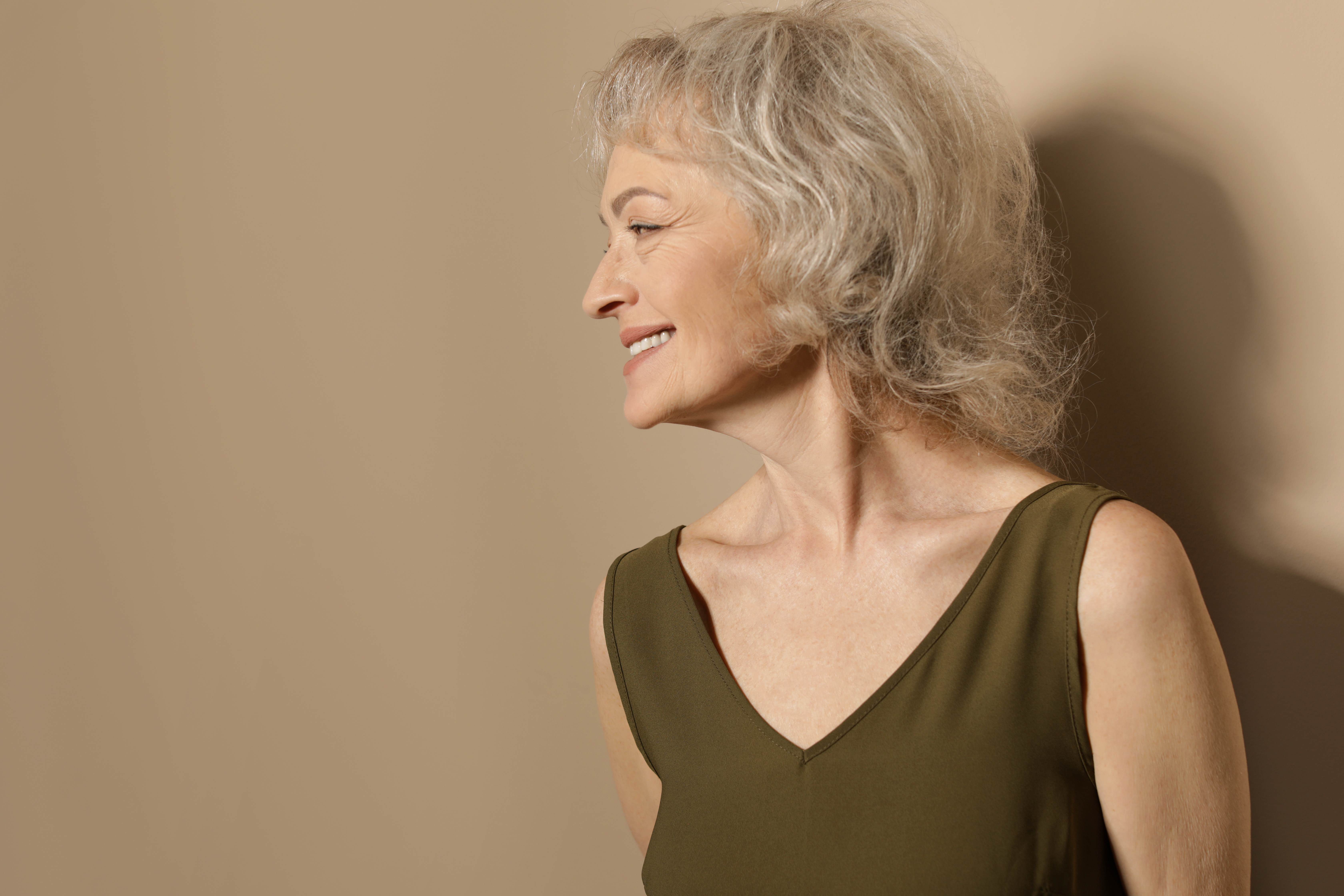 An elegant older woman in a warm beige tone background with silver-grey hair, and radiant natural skin texture, smiling softly while looking to the side.