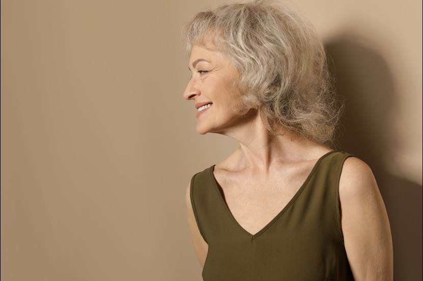 An elegant older woman in a warm beige tone background with silver-grey hair, and radiant natural skin texture, smiling softly while looking to the side.