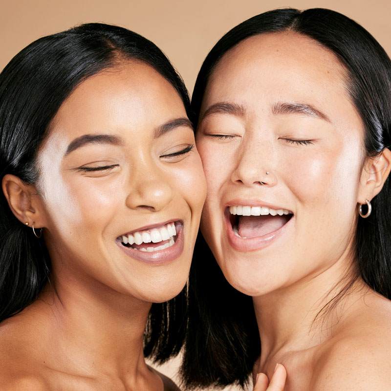 Malvallin. Two women with radiant, glowing skin share a joyful moment, laughing with eyes closed. Their expressions convey happiness and warmth on a beige background.