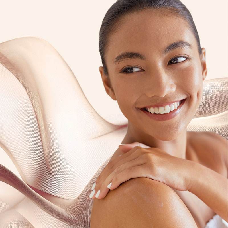 Malvallin. Smiling woman with clear, glowing skin, resting her hands on her shoulder, set against soft, flowing abstract fabric in warm beige tones that evoke skin smoothness and firmness.