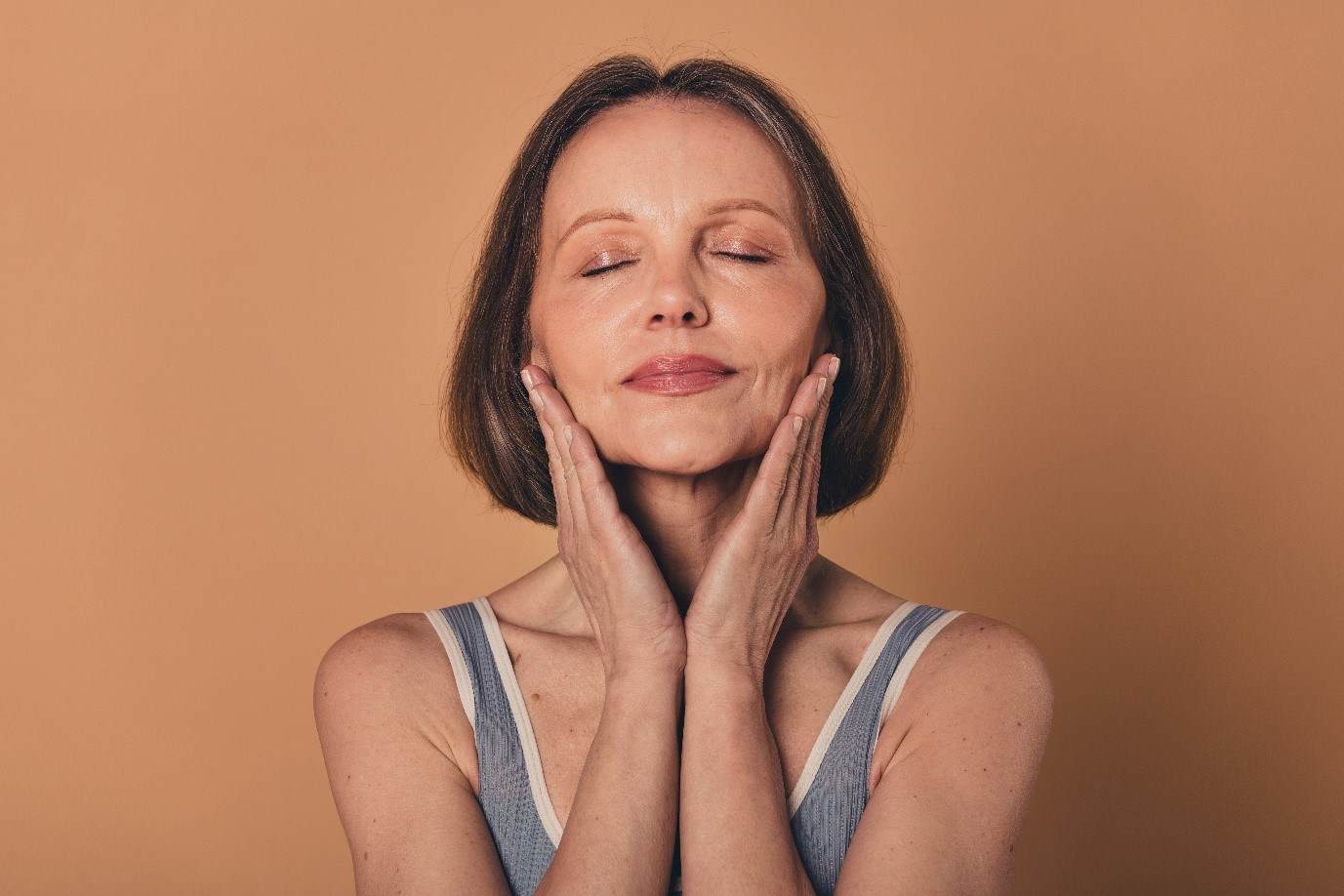 Photo of a white woman with brown hair who looks to be in her early 60s. She is applying a serum to her face with both hands. Her eyes are closed indicating how much she enjoys the refreshing serum.