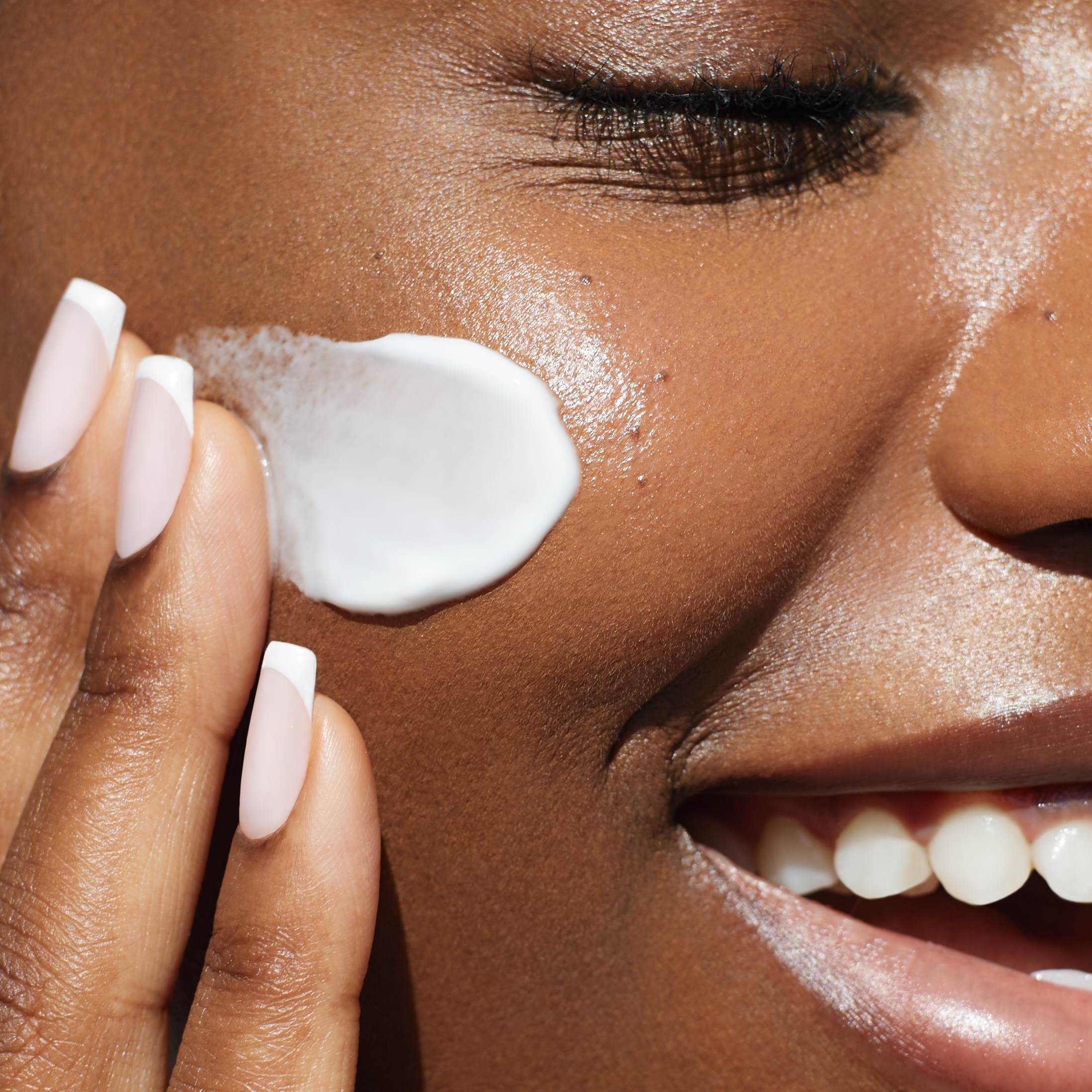 Close up on the cheek of a smiling black woman who appears to be in her 20's or 30's. She has her eyes closed and is applying a white cream. She's smiling and enjoying the sensation of applying the balm.