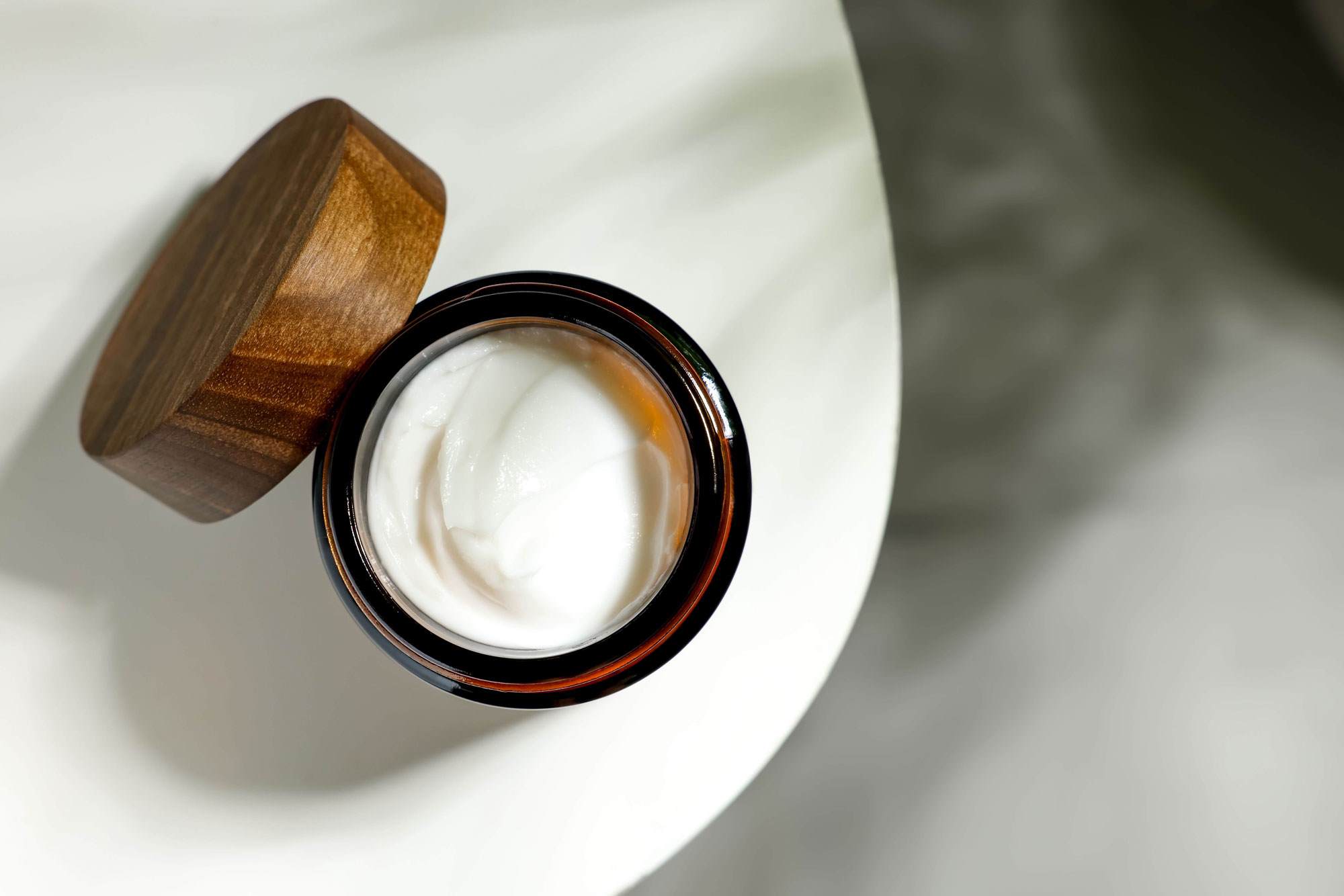 Open jar or high quality white face cream. It has a wooden lid. It is sitting on a white marble table.