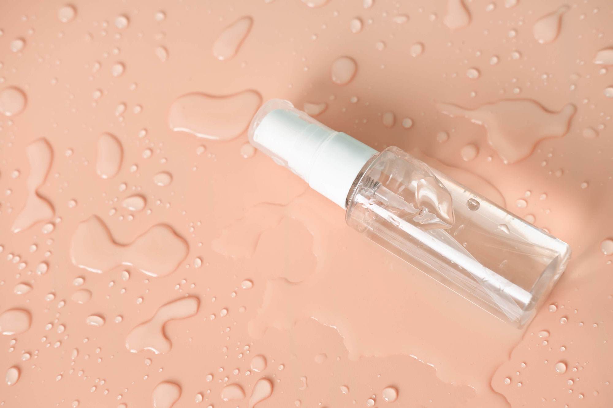 Spray bottle with a clear fluid inside on a peachy pink background There is lots of fresh water surrounding the bottle.