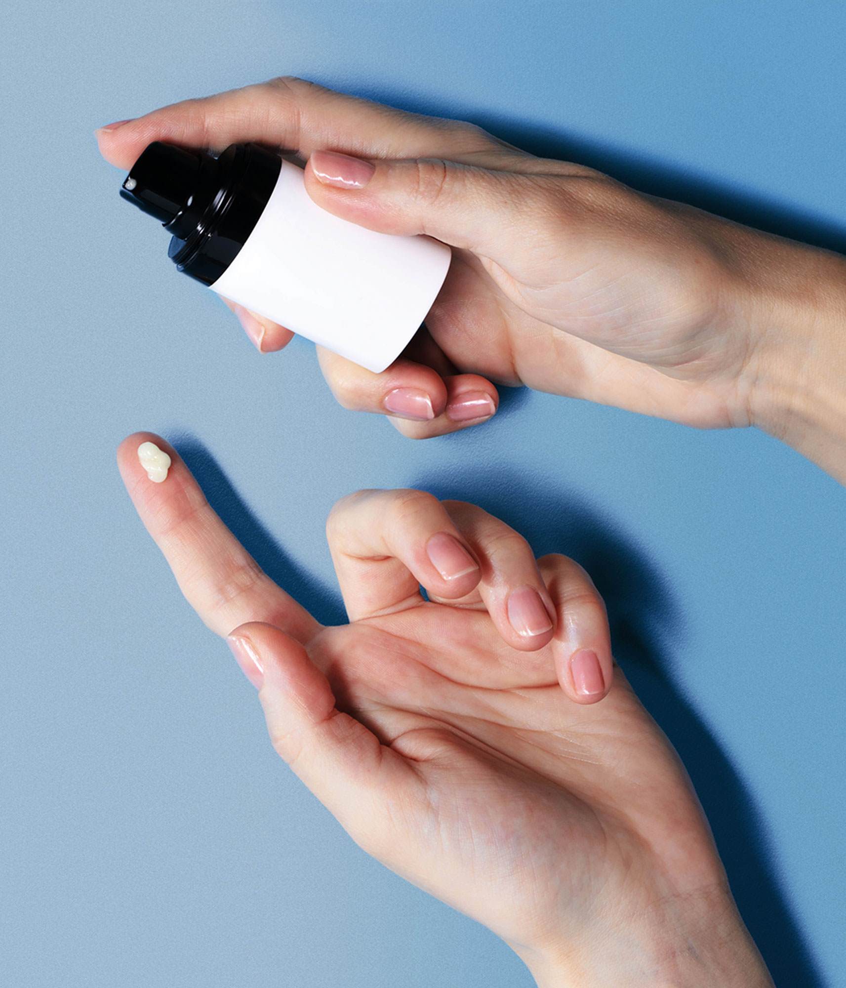 Close-up on the 2 hands of a White woman. She is holding a container of all day moisturising cream and has squirted one drop of it onto her finger.