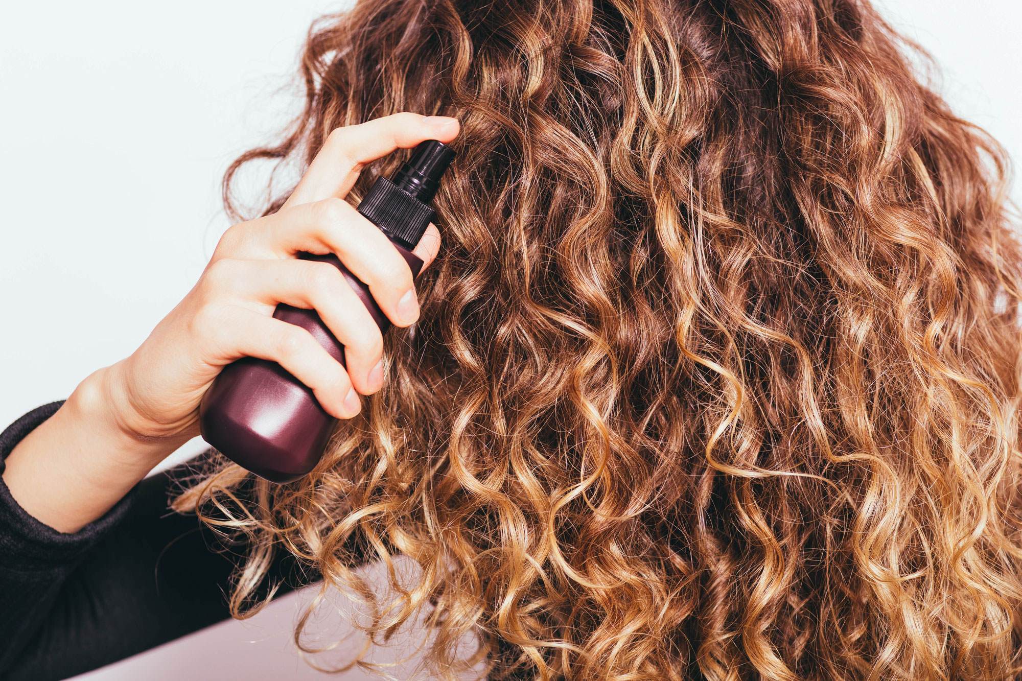 Cropped photo of a white woman with very wavy brown hair. She is spraying it with a bond-building spray.