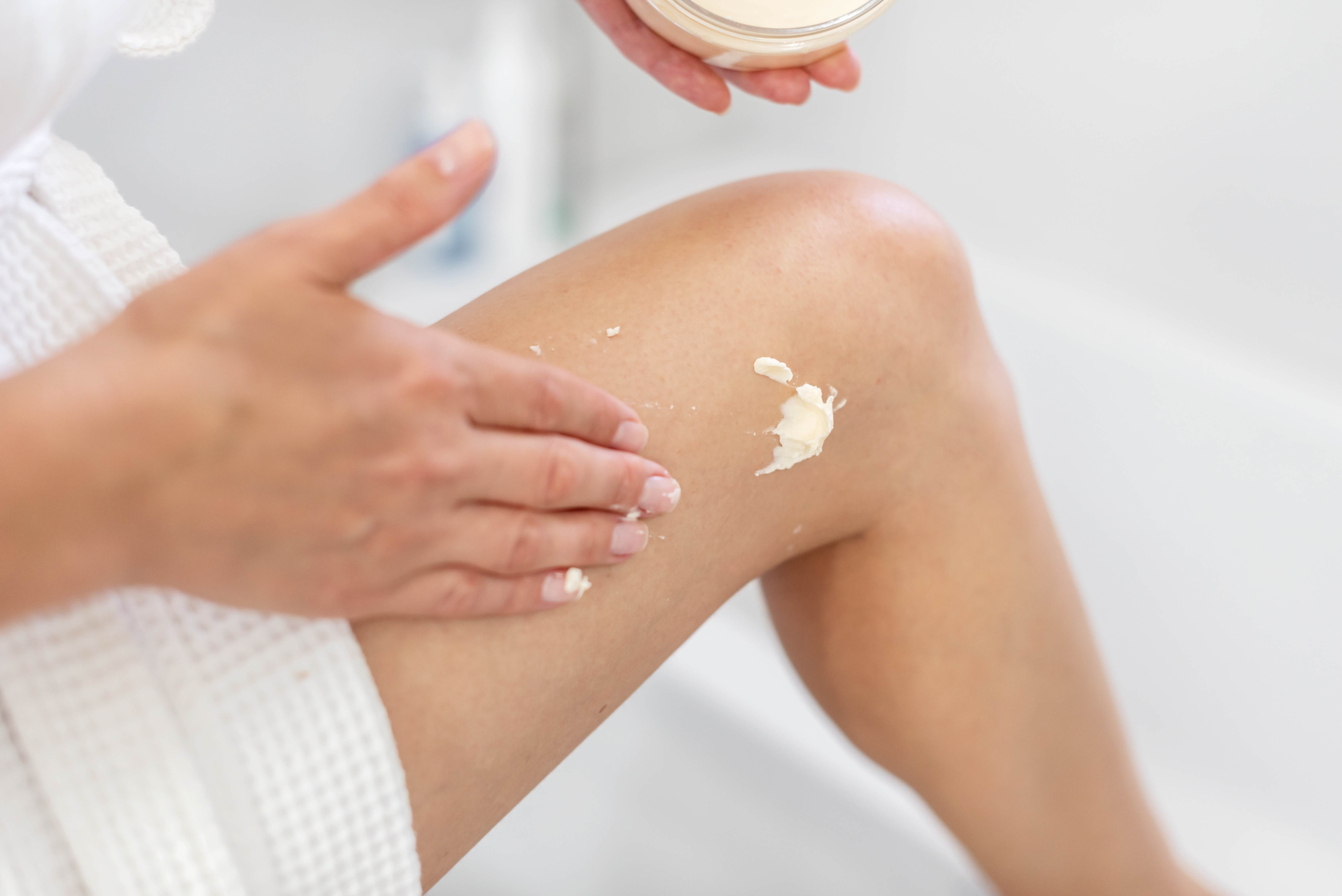Cropped image showing a White woman's knee and hand. She is applying a white cream (the care and hydrate body lotion) to her knee.