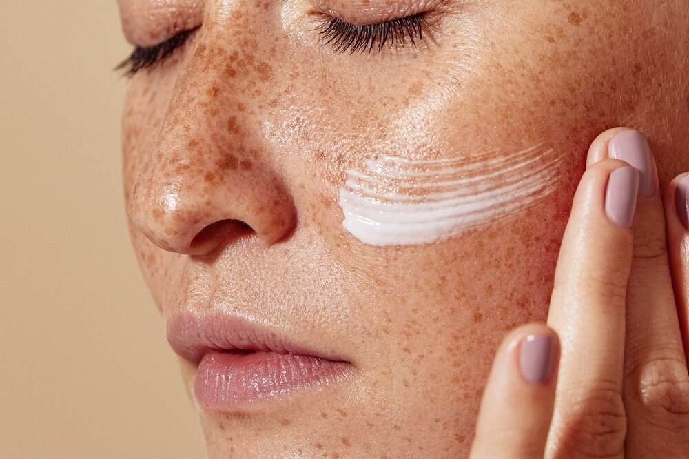 Close up on a white woman's face. She has freckles and has her eyes closed as she applies a nourishing skin cream.