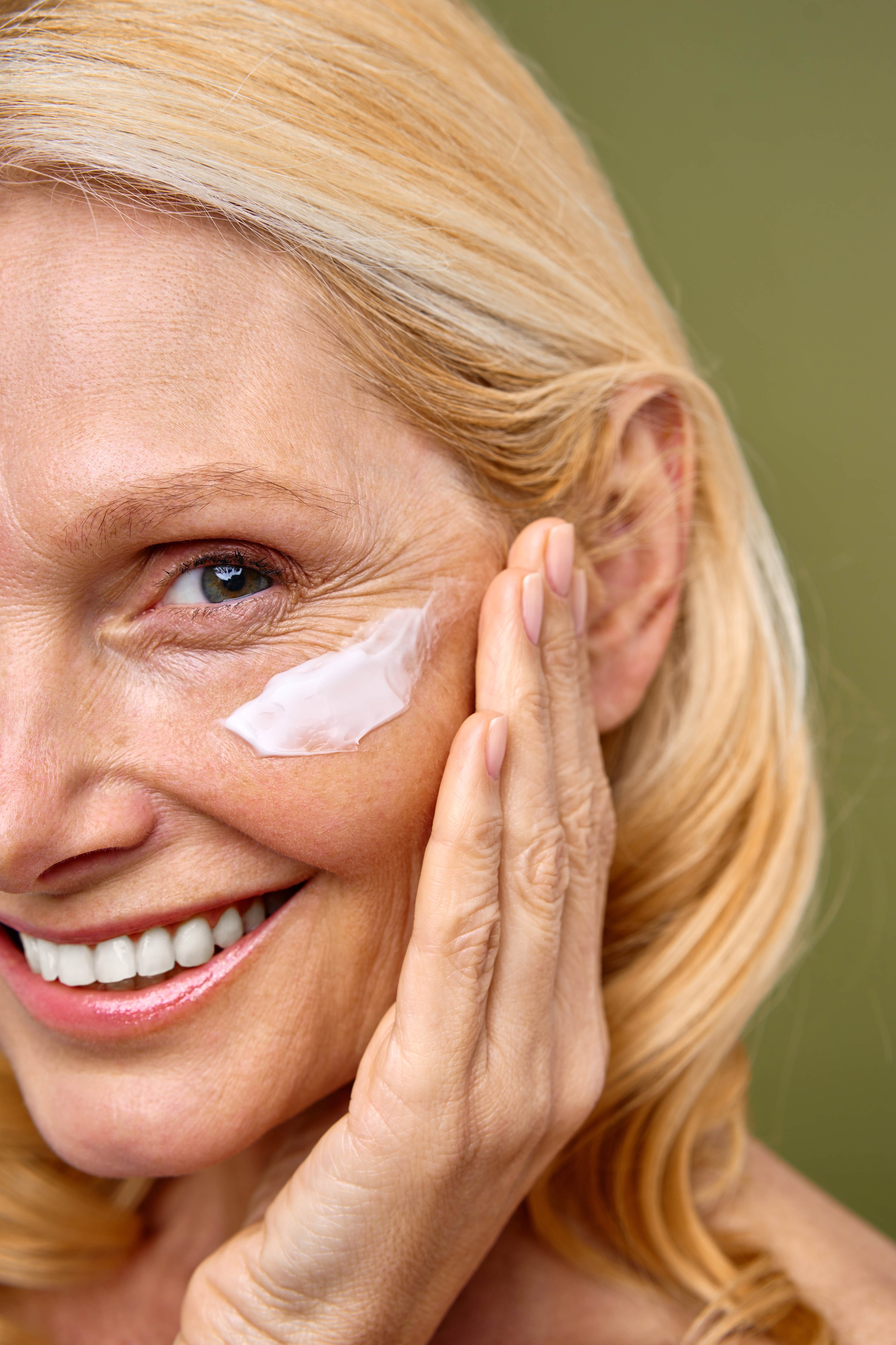 Cropped photo showing a close-up on the face of an older White woman with blonde hair. Only one eye is visible. She is smiling broadly. With one hand she is applying a white lifting serum to her face.