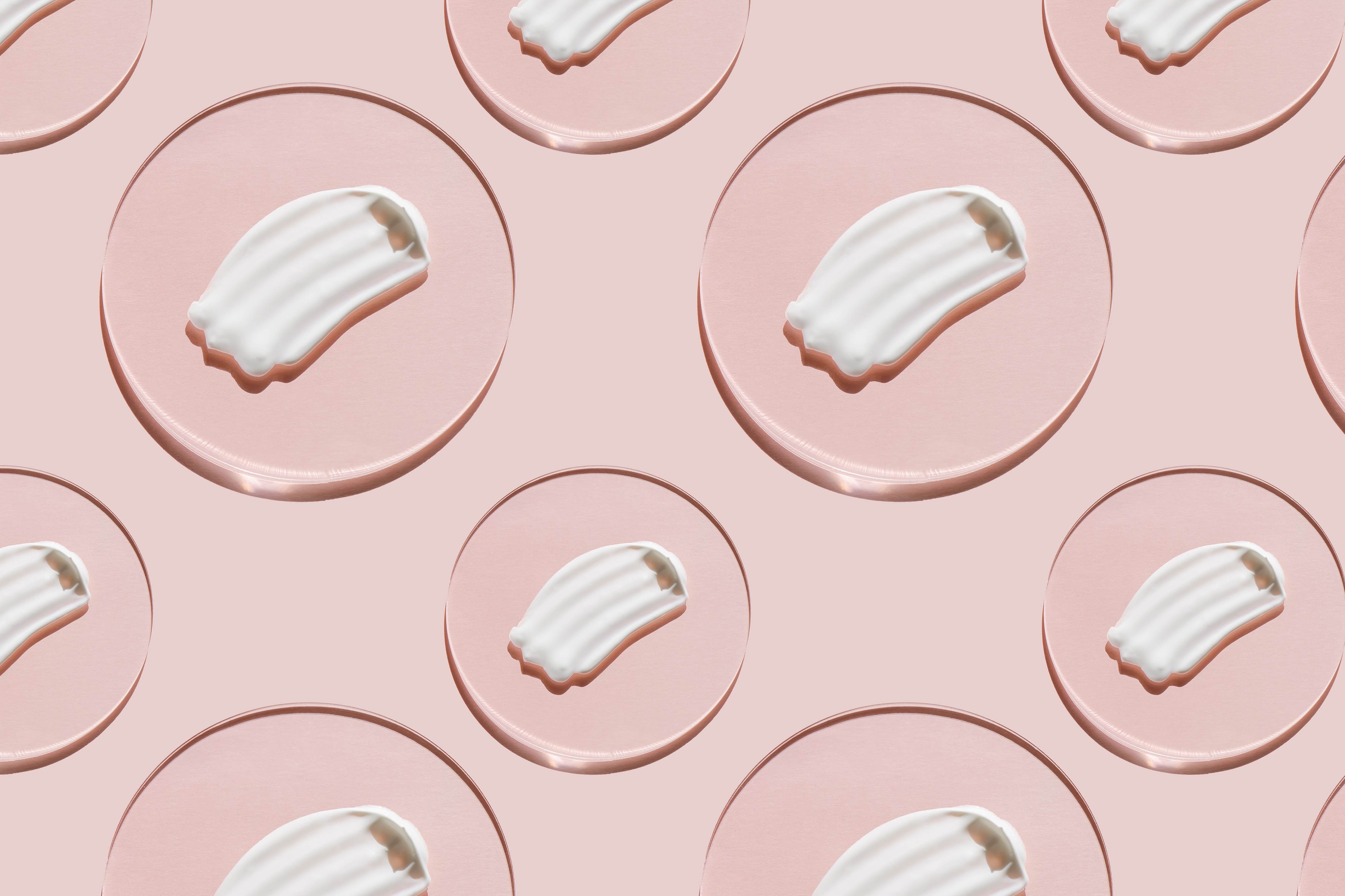 Repeating pattern of a white milky cream smear on a circular oil drop against a pink background.