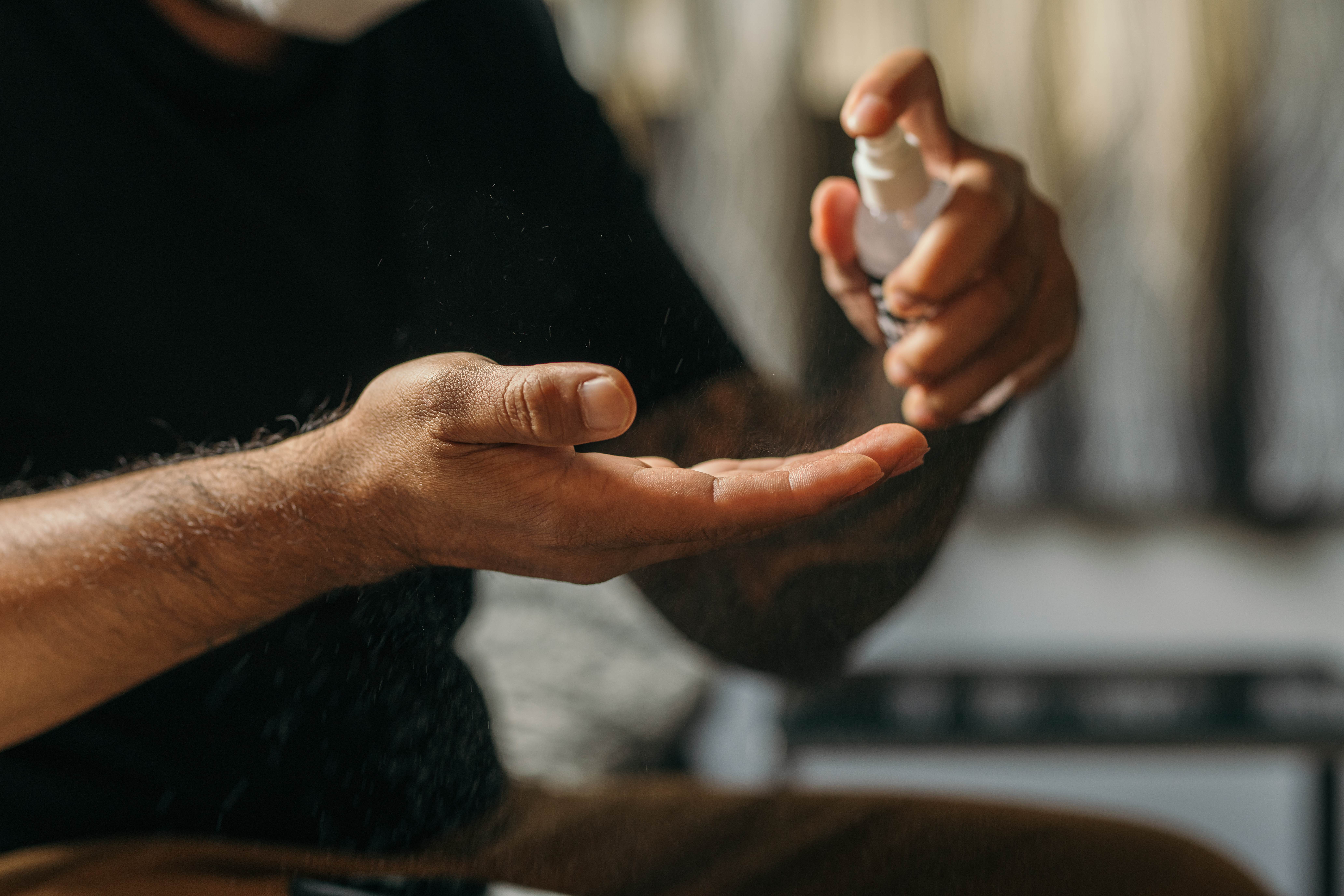 Close-up on a man's hands. The man has brown skin and is wearing a black t-shirt. He is spraying a body lotion spray into into his hands.