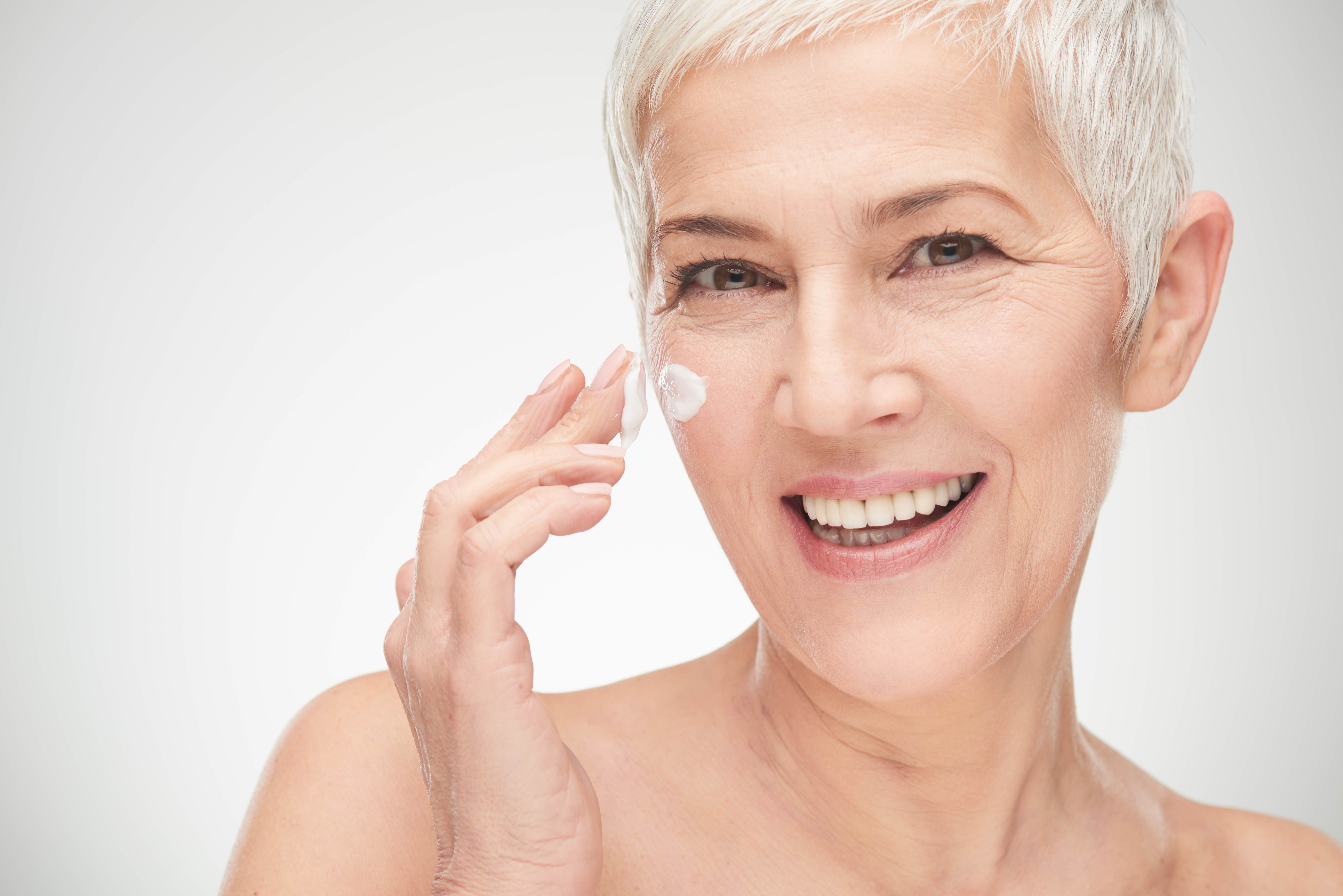 Older white woman with striking short silver hair. She is smiling, looking at the camera, and applying a white cream under her eye.