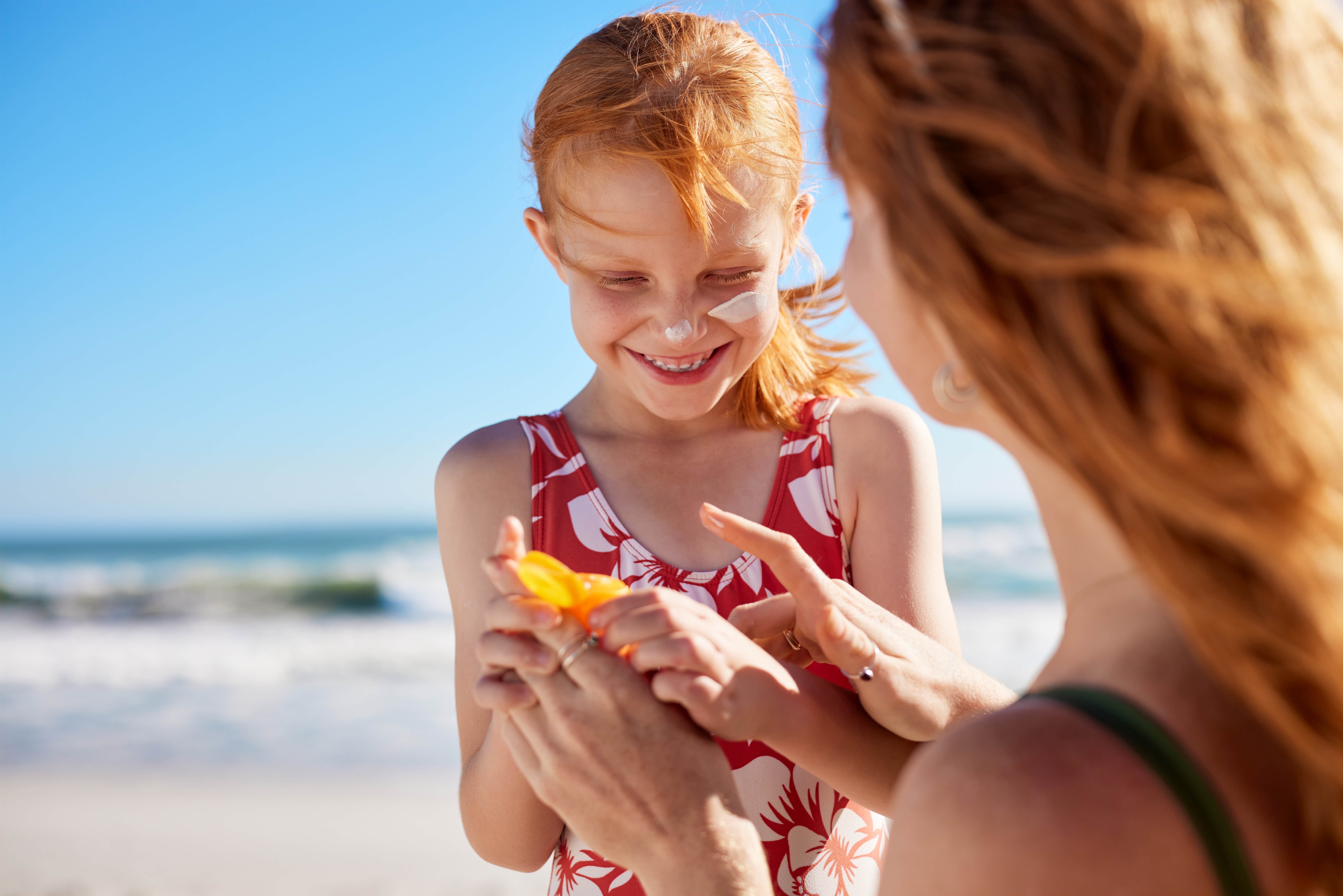 A woman with curly red hair shot from behind. she is applying sunscreen to a young white girl who also has red hair. They are at the beach. The young girl is smiling and having a fun day.