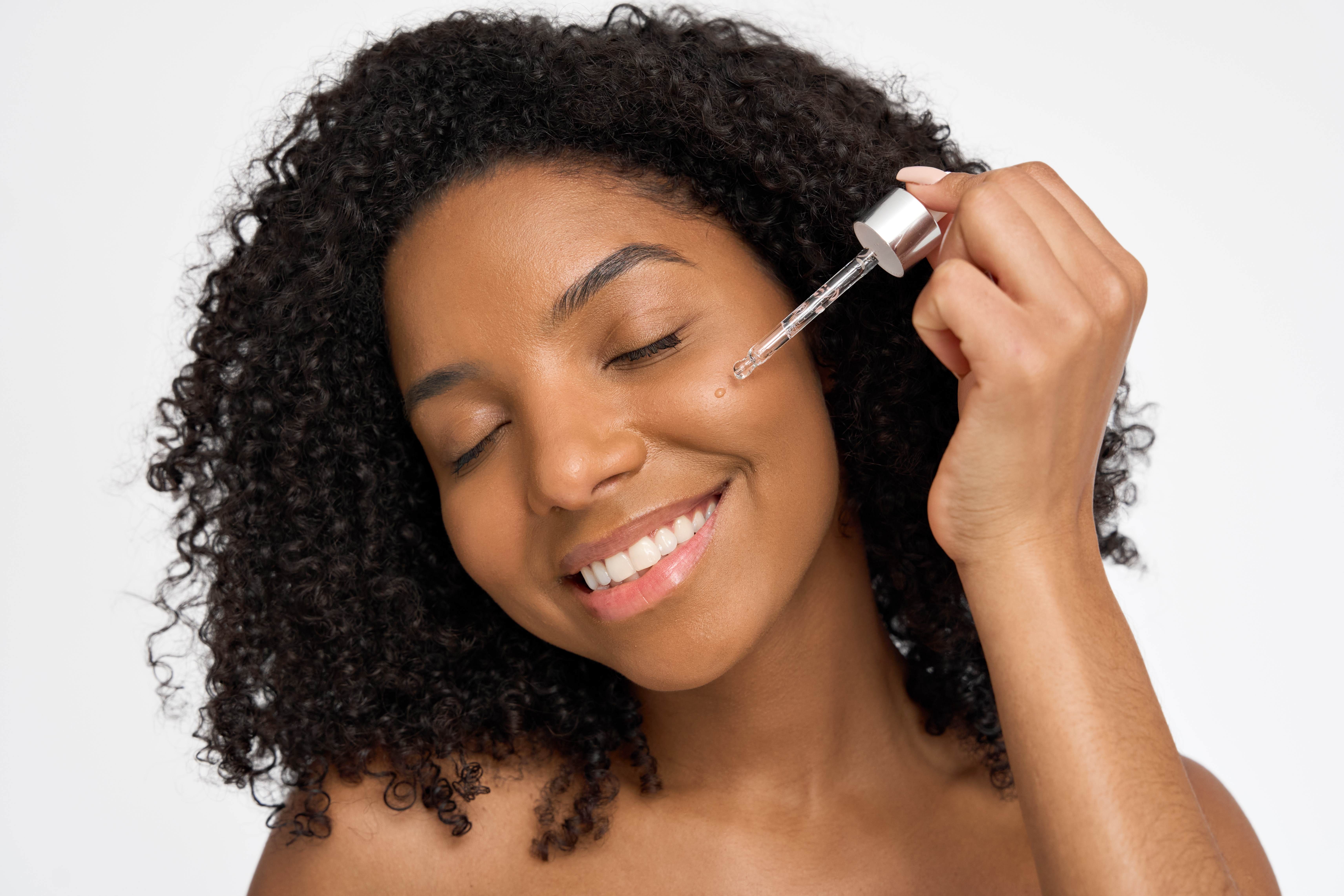Young black woman with natural black hair. She is smiling with her eyes closed and applying a serum to her cheek.