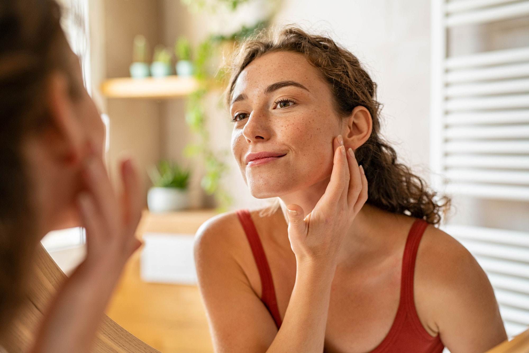 Young white woman looking in the mirror. She has curly brown hair. She is applying a morning cream and smiling.
