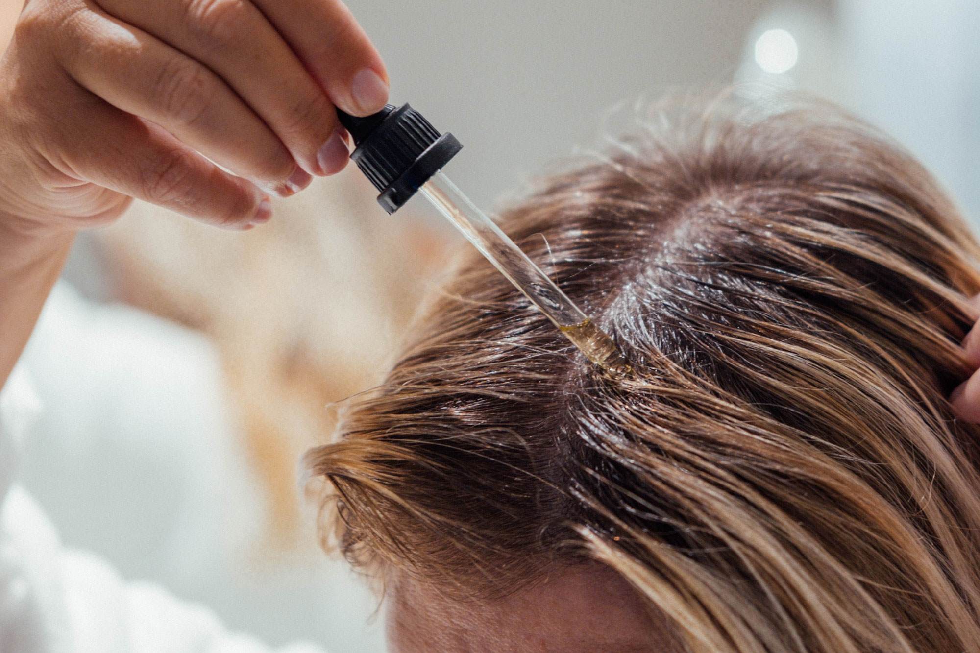 Cropped photo showing the top of a woman's head. She has brown hair and is applying a scalp soothing strand soothing serum to it with a dropper.
