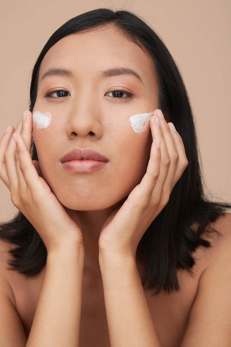 Woman of east Asian descent with light brown skin and black straight hair applying a white cream to her cheeks.