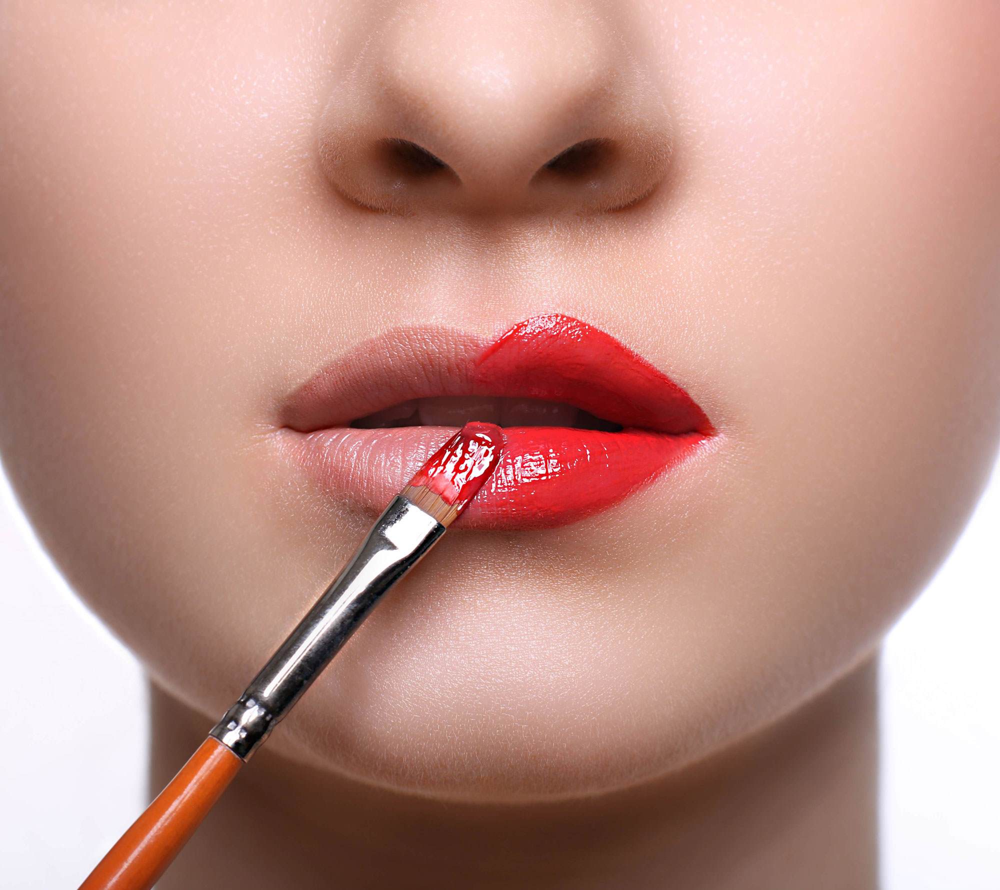Cropped close-up on a young white woman's lips as she applies ruby red lip stain to her lips using a brush.